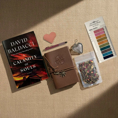 A styled flat-lay arrangement on a beige fabric background featuring a book by David Baldacci, a brown leather journal with a wrap closure, a pink heart-shaped leather book mark and tea infuser, Misty Harbor annotation tabs, and a small bag of dried flower tea.