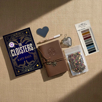A styled flat-lay featuring 'The Cloisters' book by Katy Hays, a brown leather journal with an embossed compass, a pink and white pen, a heart-shaped leather bookmark and tea infuser, Gothic Romance annotation tabs from Fehmerling Books and a bag of dried flower tea, all arranged on a beige linen background with diagonal shadows.