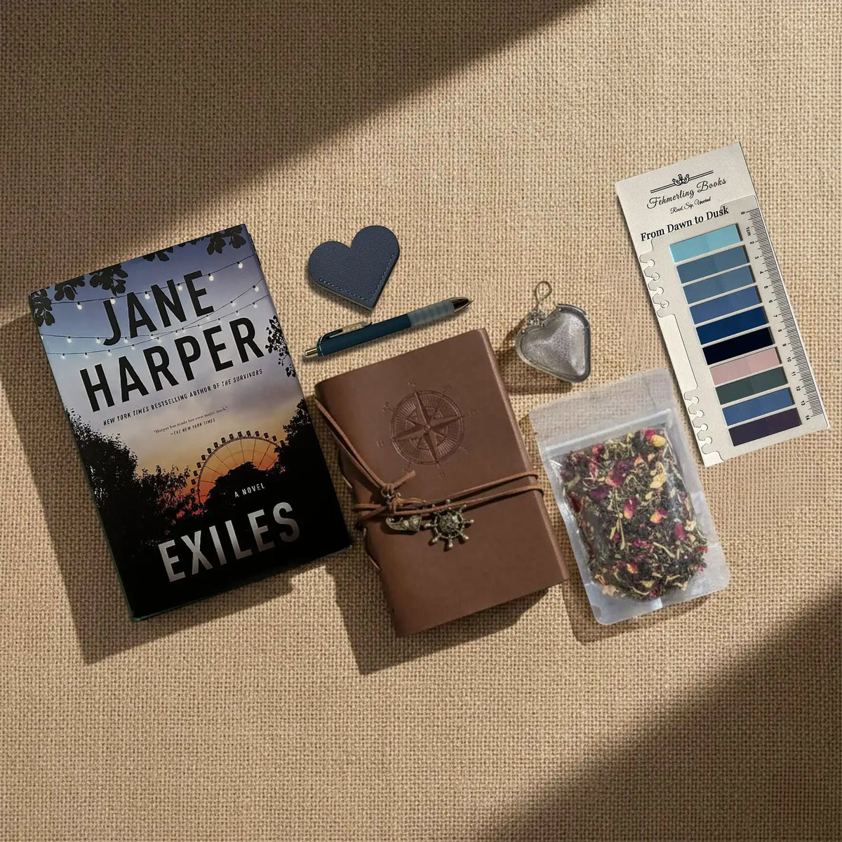 A styled flat-lay of a Jane Harper book titled 'Exiles' alongside a brown leather journal, a heart-shaped tea infuser and leather bookmark,  a tea bag packet, and From Dawn to Dusk annotation tabs, all arranged on a beige linen background with diagonal shadows.
