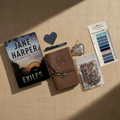 A styled flat-lay of a Jane Harper book titled 'Exiles' alongside a brown leather journal, a heart-shaped tea infuser and leather bookmark,  a tea bag packet, and From Dawn to Dusk annotation tabs, all arranged on a beige linen background with diagonal shadows.