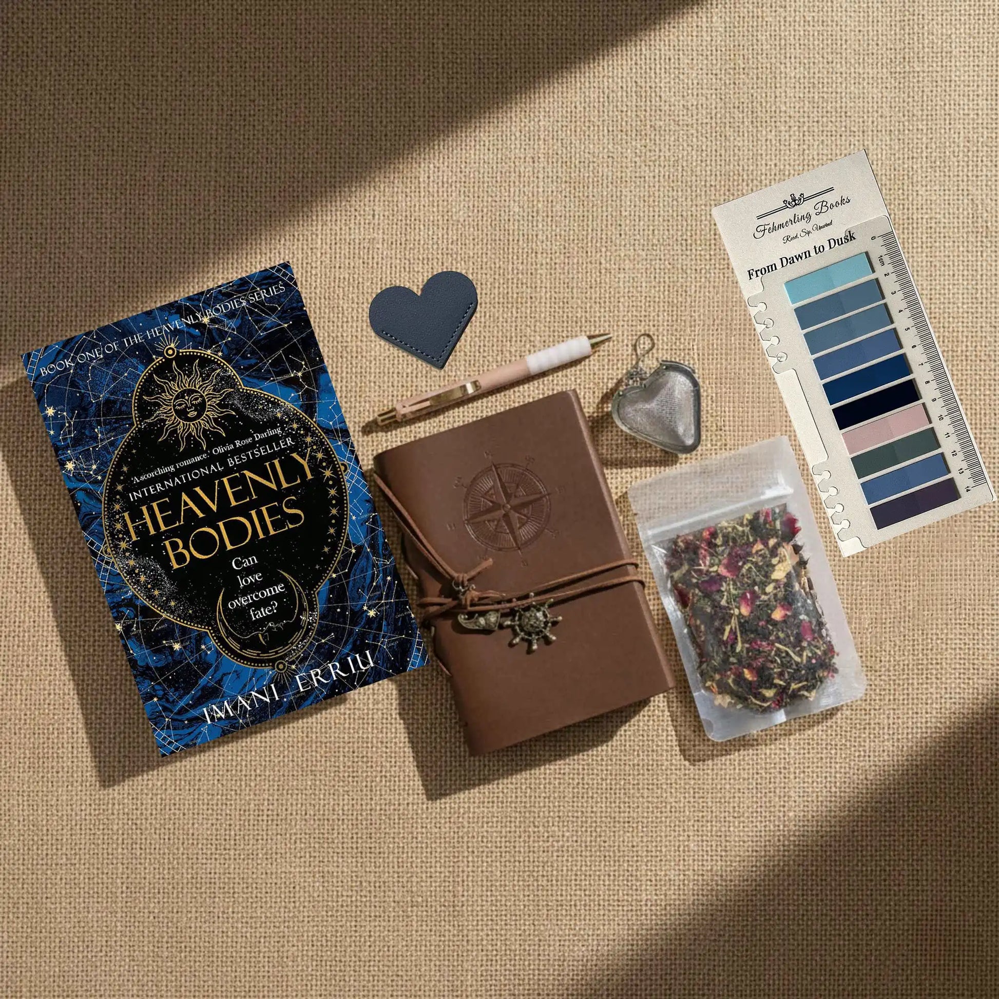 A styled flat-lay on a beige linen background featuring a book titled 'Heavenly Bodies' by Imani Erriu, a brown leather journal with an embossed compass, a pen, a heart-shaped tea infuser, From Dawn to Dusk annotation tabs, and a package of herbal tea.