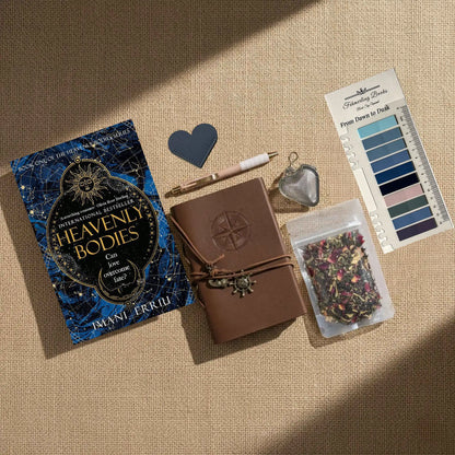 A styled flat-lay on a beige linen background featuring a book titled 'Heavenly Bodies' by Imani Erriu, a brown leather journal with an embossed compass, a pen, a heart-shaped tea infuser, From Dawn to Dusk annotation tabs, and a package of herbal tea.