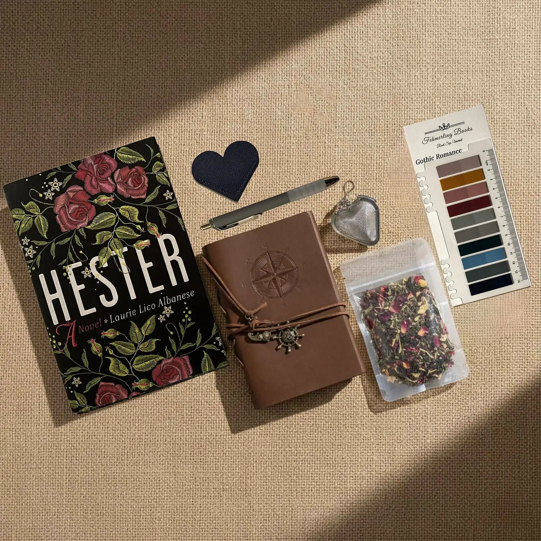 A styled flat-lay of Hester journaling supplies on a beige linen background, including  a brown leather-bound journal with an embossed compass, Gothic Romance annotation tabs from Fehmerling Books, dried flower tea, a hear-shaped leather bookmark and tea infuser and a bag of herbal tea.