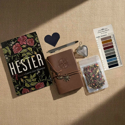 A styled flat-lay of Hester journaling supplies on a beige linen background, including  a brown leather-bound journal with an embossed compass, Gothic Romance annotation tabs from Fehmerling Books, dried flower tea, a hear-shaped leather bookmark and tea infuser and a bag of herbal tea.