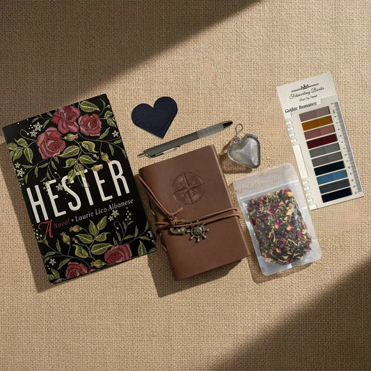 A styled flat-lay of Hester journaling supplies on a beige linen background, including  a brown leather-bound journal with an embossed compass, Gothic Romance annotation tabs from Fehmerling Books, dried flower tea, a hear-shaped leather bookmark and tea infuser and a bag of herbal tea.