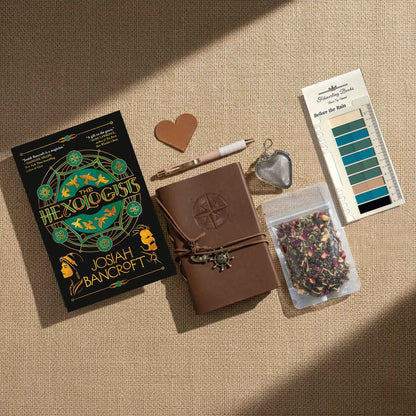 A styled flat-lay of The Hexologists by Josiah Bancroft on a beige linen background. The kit includes a brown leather-bound notebook with a compass embossing, a heart-shaped leather bookmark and tea infuser, a pen, a clear bag of dried flower tea, and Before the Rain annotation tabs from Fehmerling Books.