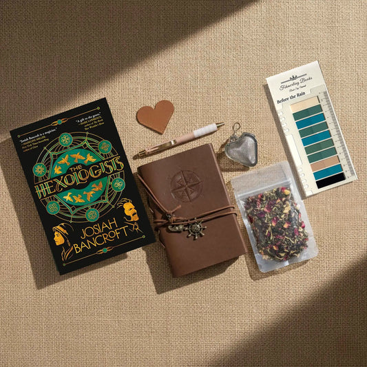 A styled flat-lay of The Hexologists by Josiah Bancroft on a beige linen background. The kit includes a brown leather-bound notebook with a compass embossing, a heart-shaped leather bookmark and tea infuser, a pen, a clear bag of dried flower tea, and Before the Rain annotation tabs from Fehmerling Books.