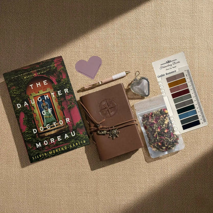 A styled flat-lay of a cozy reading-themed stationery and book accessory set on a beige linen background. The set includes a copy of the book 'The Daughter of Doctor Moreau' by Silvia Moreno-Garcia, a brown leather-bound journal with an embossed compass, a white and rose gold pen, a heart-shaped leather bookmark and tea infuser, Gothic Romance annotation tabs from Fehmerling Books, and a small bag of dried flower tea.