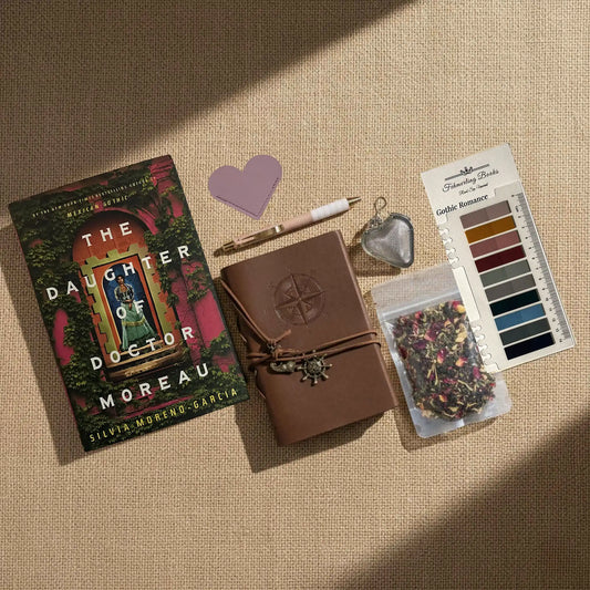 A styled flat-lay of a cozy reading-themed stationery and book accessory set on a beige linen background. The set includes a copy of the book 'The Daughter of Doctor Moreau' by Silvia Moreno-Garcia, a brown leather-bound journal with an embossed compass, a white and rose gold pen, a heart-shaped leather bookmark and tea infuser, Gothic Romance annotation tabs from Fehmerling Books, and a small bag of dried flower tea.