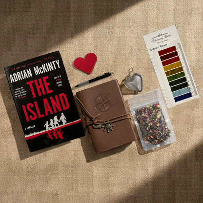 A styled flat-lay on a beige burlap background featuring a copy of the book 'The Island' by Adrian McKinty, a brown leather journal with an embossed compass, a heart-shaped leather book mark and tea infuser, a tea blend packet, and Autumn Woods annotation tabs from Fehmerling Books.