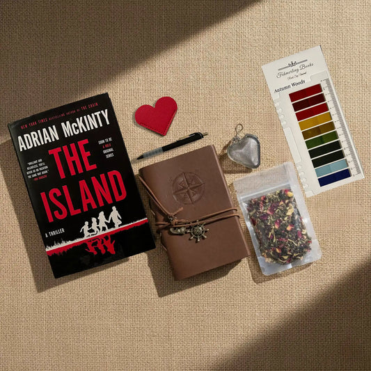 A styled flat-lay on a beige burlap background featuring a copy of the book 'The Island' by Adrian McKinty, a brown leather journal with an embossed compass, a heart-shaped leather book mark and tea infuser, a tea blend packet, and Autumn Woods annotation tabs from Fehmerling Books.