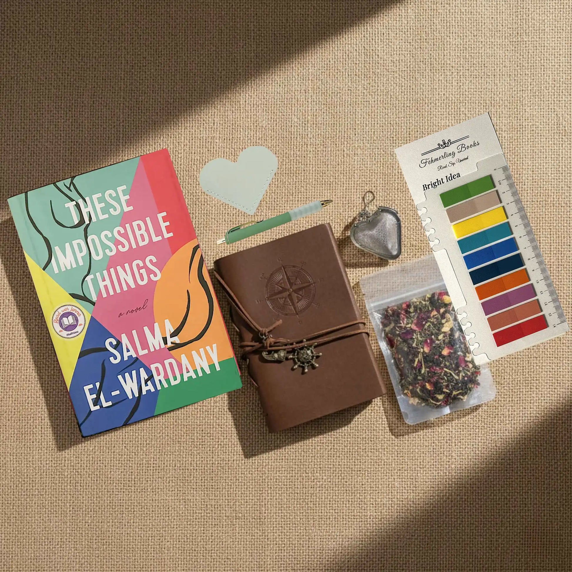 A styled flat-lay on a beige burlap background featuring a book titled 'These Impossible Things' by Salma El-Wardany, a brown leather journal with a compass embossing, a heart-shaped leather book mark and tea infuser, a packet of herbal tea, and Bright Idea annotation tabs.