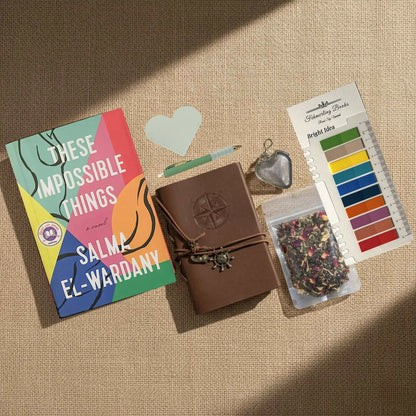 A styled flat-lay on a beige burlap background featuring a book titled 'These Impossible Things' by Salma El-Wardany, a brown leather journal with a compass embossing, a heart-shaped leather book mark and tea infuser, a packet of herbal tea, and Bright Idea annotation tabs.