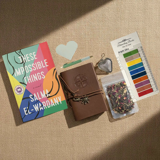 A styled flat-lay on a beige burlap background featuring a book titled 'These Impossible Things' by Salma El-Wardany, a brown leather journal with a compass embossing, a heart-shaped leather book mark and tea infuser, a packet of herbal tea, and Bright Idea annotation tabs.