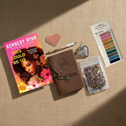 A styled flat-lay of a book and matching accessories on a beige fabric background with diagonal shadows. The set includes the book 'This Could Be Us' by Kennedy Ryan, a brown leather-bound journal with an embossed compass and wrap closure, a pink heart-shaped leather bookmark and tea infuser, a pen, Misty Harbor annotation tabs, and a small bag of dried flower tea.